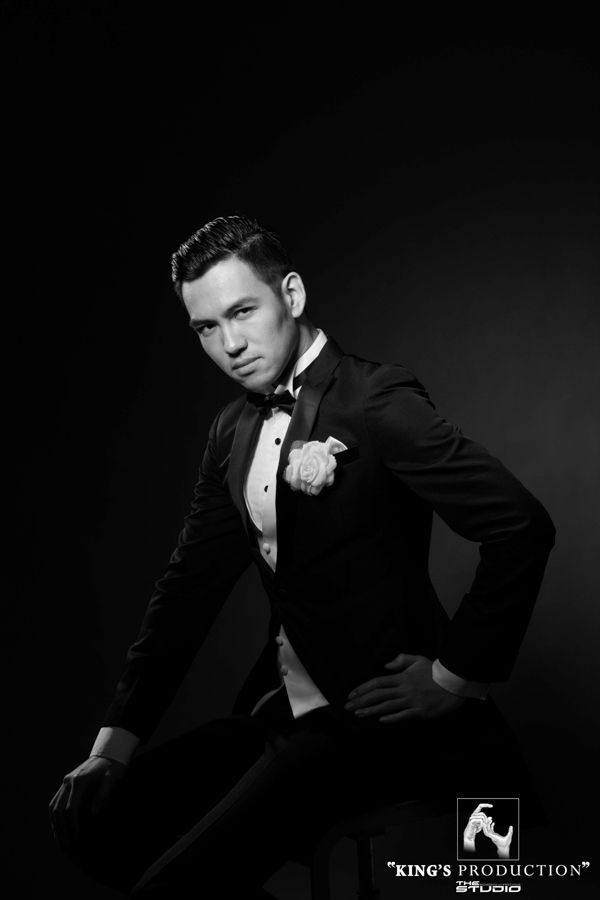 身穿燕尾服的男子坐著，帶著微笑地看著鏡頭，攝影棚燈光下。