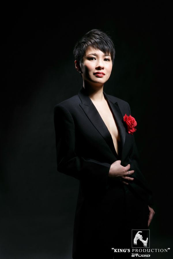 身穿黑色西裝、戴著紅花、留著短髮、塗著紅色口紅的女人在深色背景下擺姿勢。