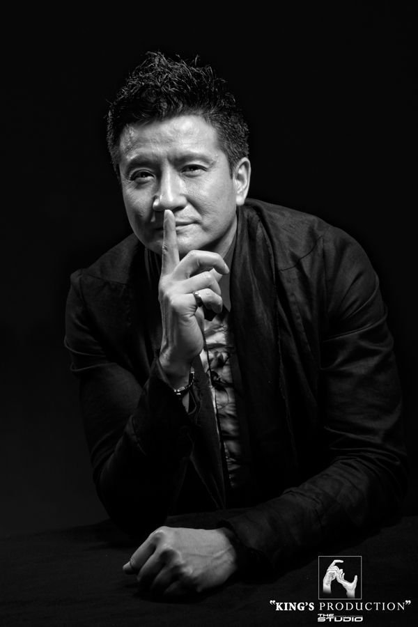 亞洲男子身穿深色西裝外套，手指放在嘴唇上，背景為黑色。