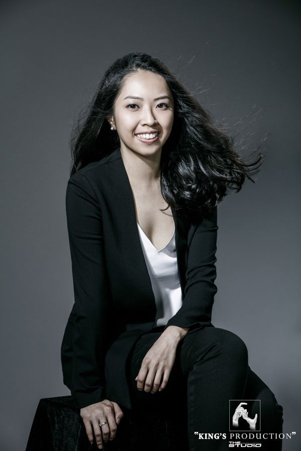 身穿黑色西裝外套和褲子的女人，帶著微笑，坐在灰色背景前。