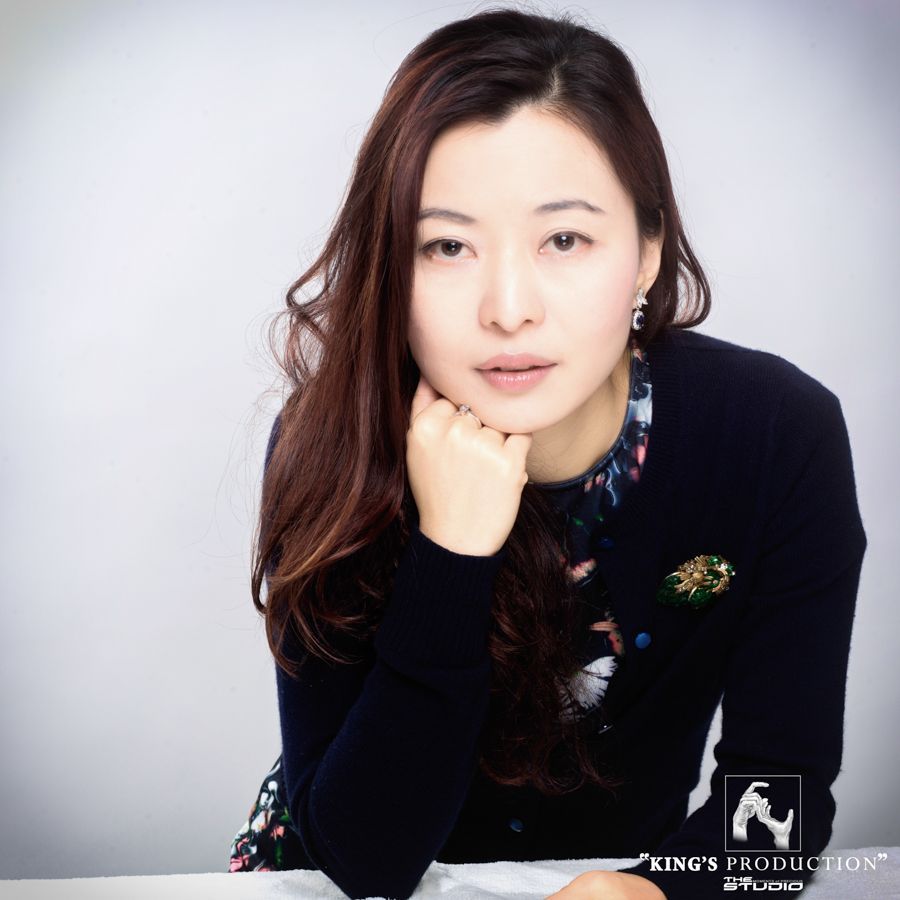 一名女子留著長長的黑髮，手托著下巴，身穿深色毛衣，裡面襯著花襯衫，坐在工作室裡。