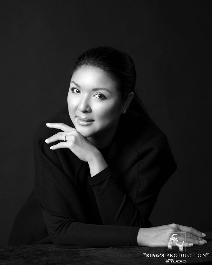 身穿黑色洋裝的女子，雙臂休息，在黑暗的背景下對著鏡頭微笑。