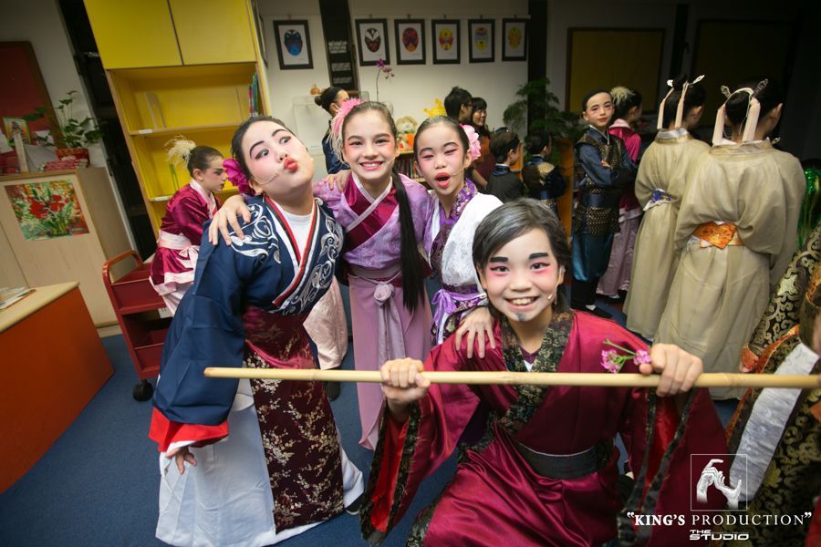身著中國戲曲服裝的孩子們在後台擺姿勢，面帶微笑，手裡拿著道具。