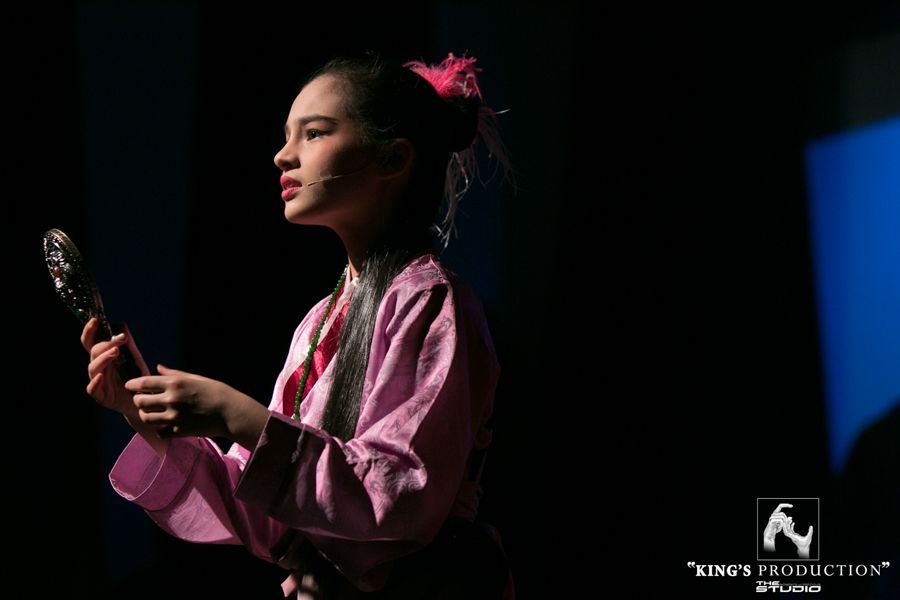 身穿粉紅色服裝的年輕女演員，手拿鏡子，站在黑暗的舞台上。