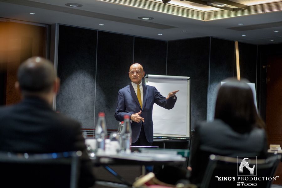 一位西裝革履的男士在桌旁做演講。觀眾席中有兩人。會議室場景。