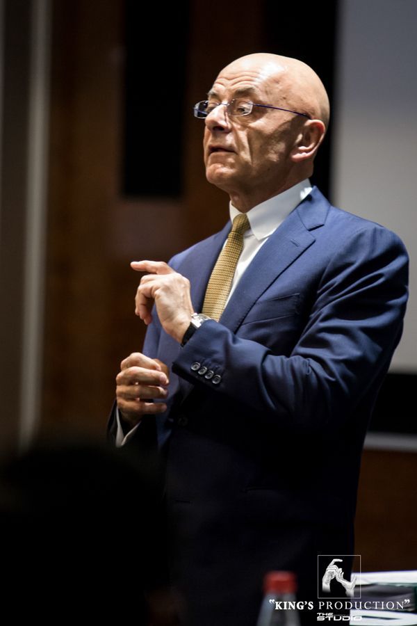 身穿藍色西裝、戴眼鏡和領帶的男子在室內演講，指著什麼。