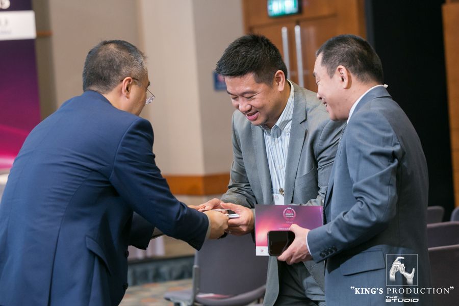 三位男士在一場活動中握手。他們身處一間有座位的房間裡。右邊的男士手裡拿著一部手機和一本小冊子。