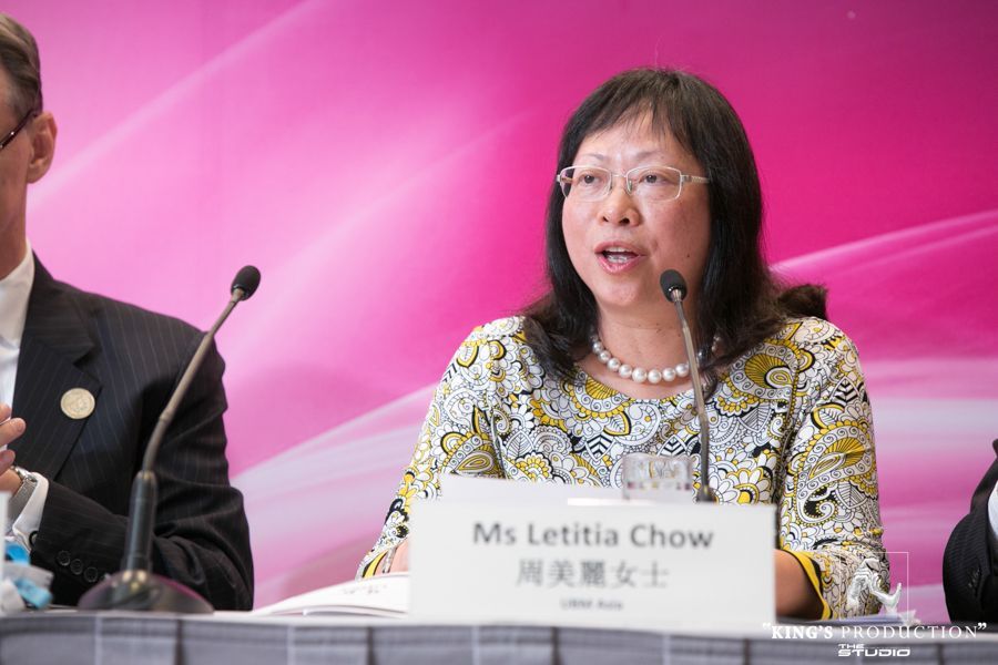 週女士在會議上發言。她戴著眼鏡，穿著有圖案的上衣，戴著珍珠項鍊，背景是粉紅色的。