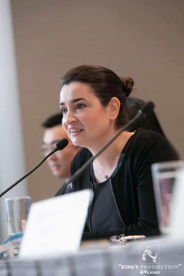 一位女士在小組討論會上發言，深色頭髮盤成髮髻，穿著黑色衣服，拿著麥克風，背景明亮。