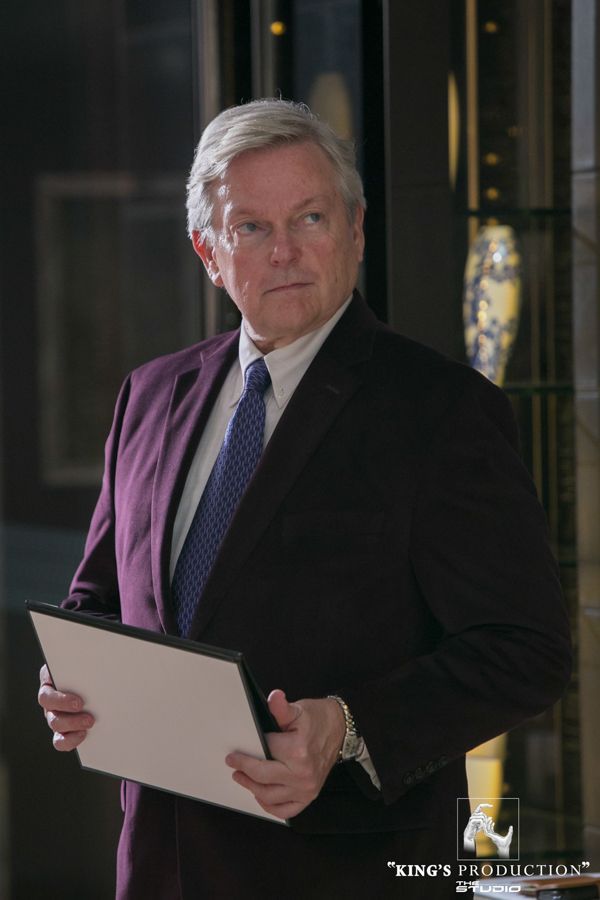 身穿紫色西裝的男子手持文件，若有所思。室內場景。