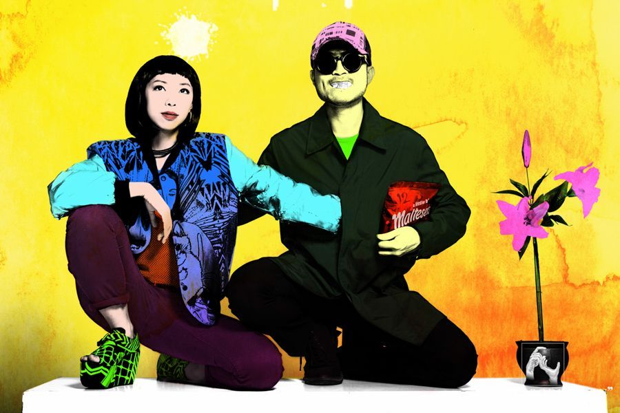 兩個身著鮮豔服裝的人在黃色背景前擺姿勢。女人跪著，男人坐著，手裡拿著一個袋子。