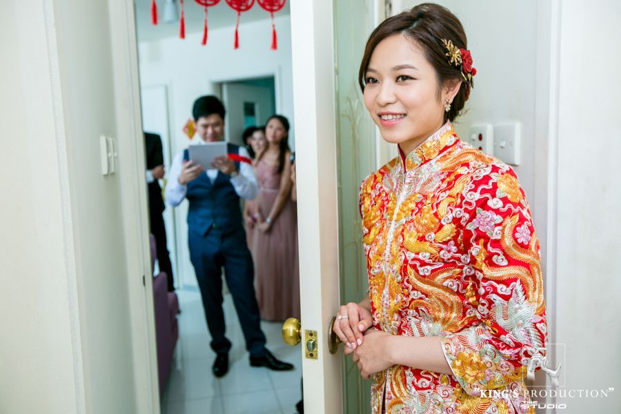 新娘身穿紅色和金色的傳統服飾，在門口微笑；客人在背景中，房間裝飾一新。