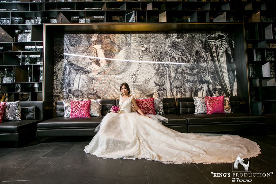 穿著白色禮服的新娘坐在黑色沙發上，身邊放著粉紅色枕頭，前面是壁畫和書架。