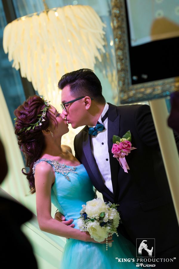 新娘和新郎接吻，他穿著燕尾服，她穿著綠松石色禮服，手捧鮮花，室內婚禮場景。