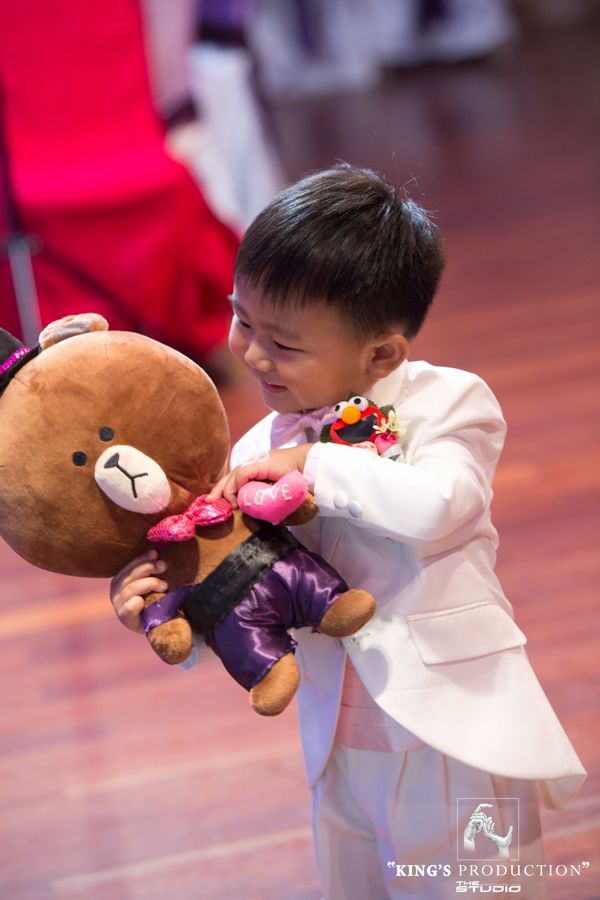 活動中，穿著白色西裝的小男孩微笑著擁抱一隻棕熊娃娃。