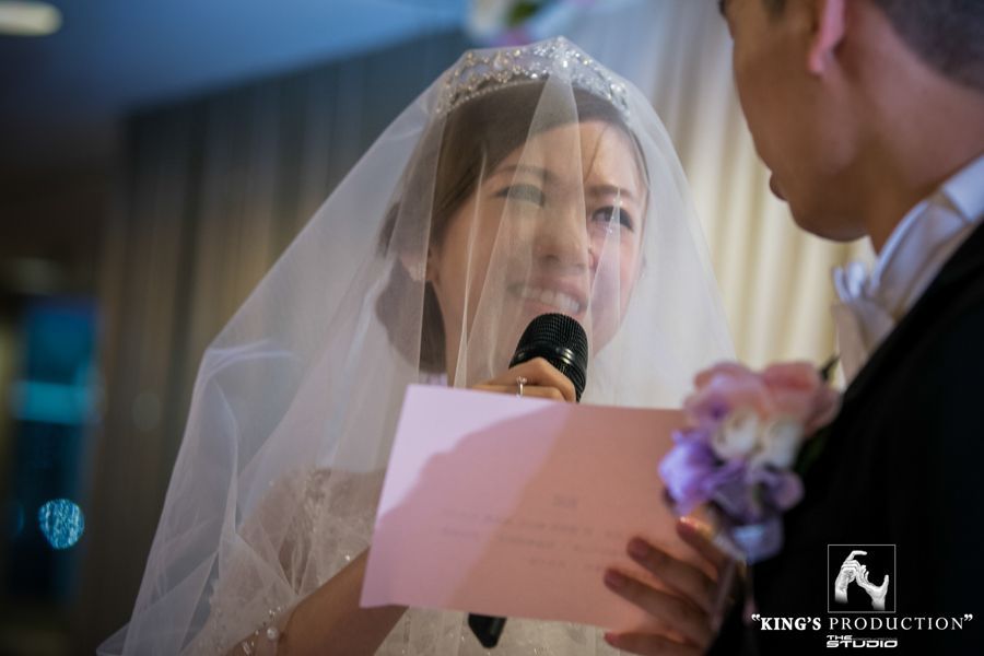 婚禮上，新娘戴著面紗，手拿麥克風和卡片，看著新郎。