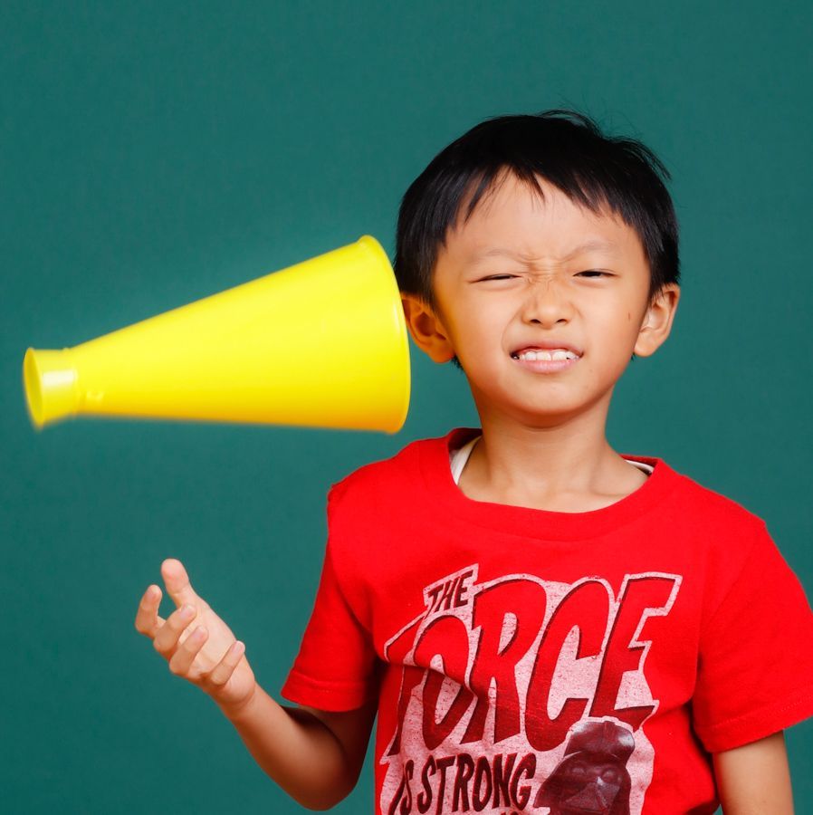 男孩瞇著眼睛，耳朵附近有黃色擴音器，穿著紅色襯衫，背景是綠色。