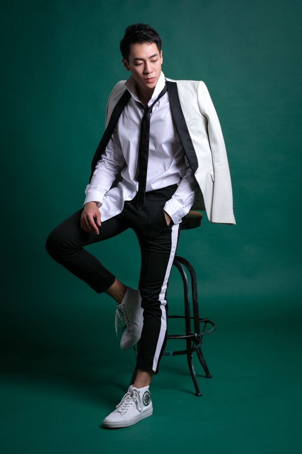 一名男子身穿白襯衫、黑褲（白色條紋），坐在凳子上，外套搭在肩上，低頭看著前方。背景為綠色。