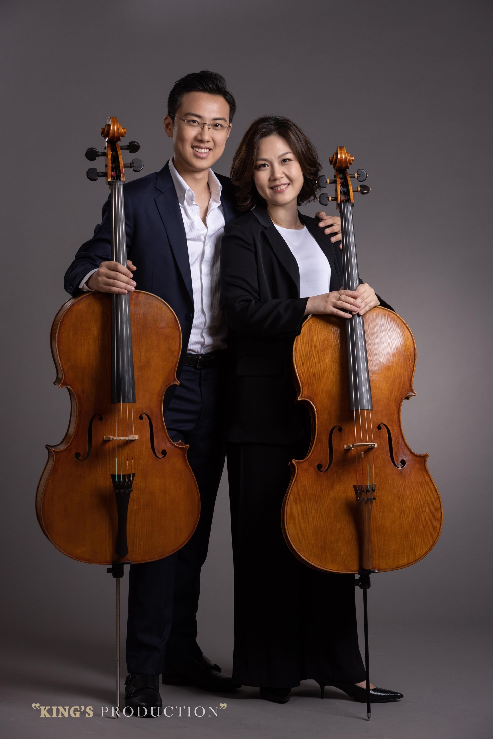 兩位大提琴手，一男一女，在灰色背景下微笑著，手裡拿著大提琴。