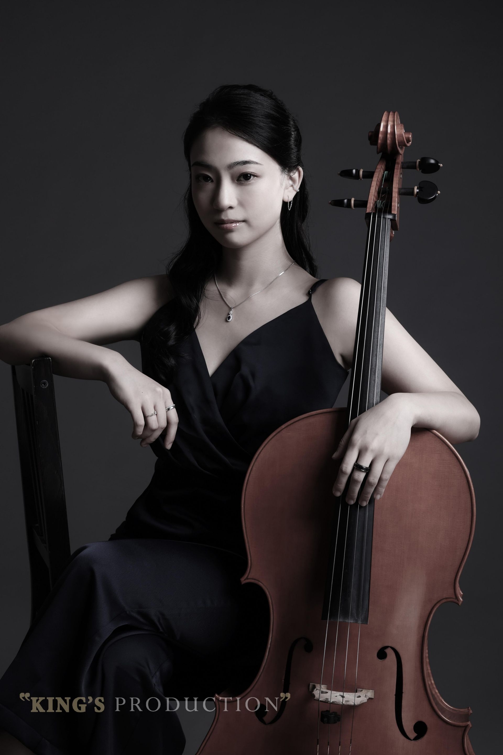 身穿深色連身裙、手持大提琴的女子坐在灰色背景前。