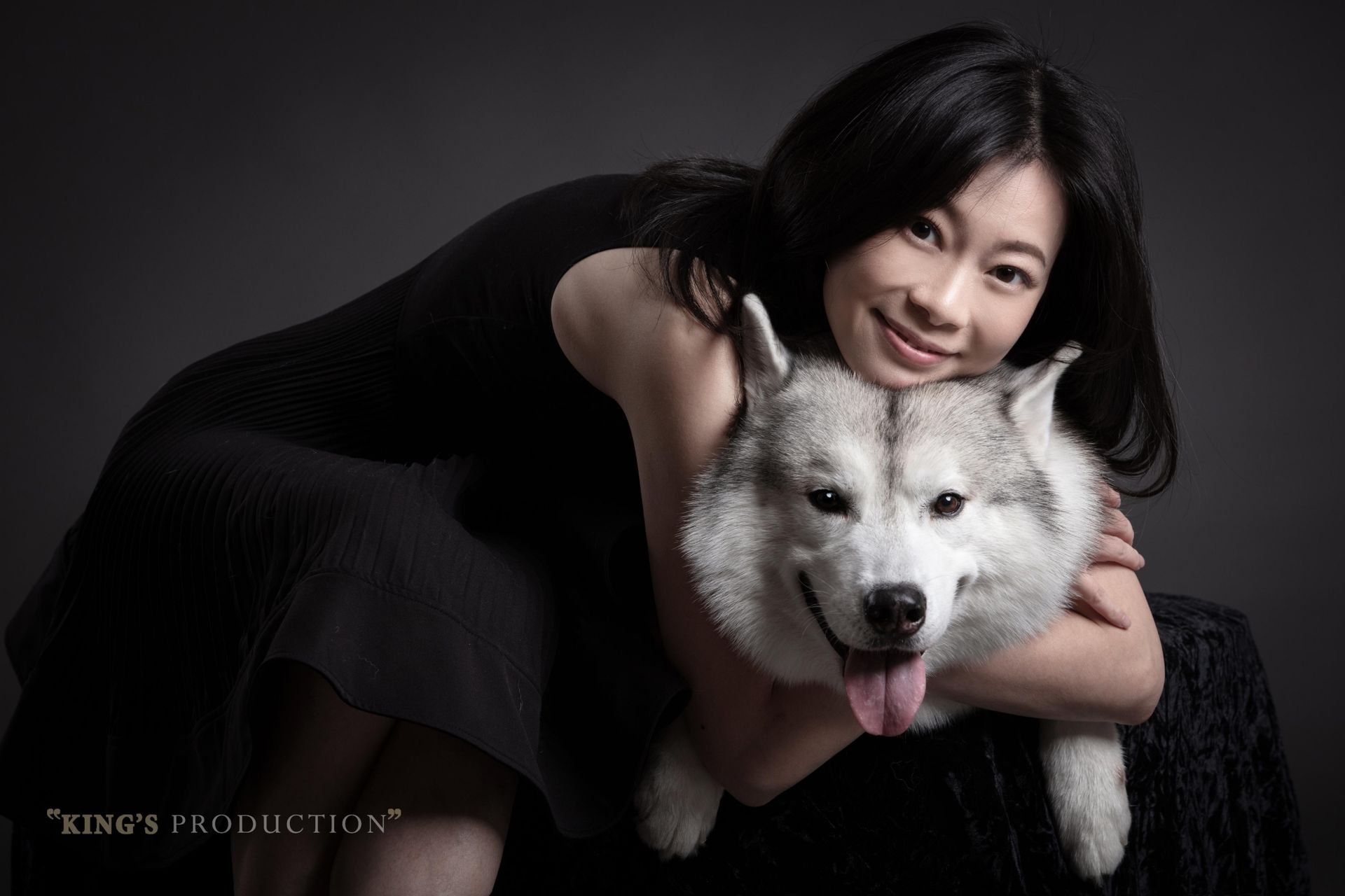 身穿黑色連身裙的女子擁抱著一隻毛茸茸的灰白色西伯利亞哈士奇，兩人都面帶微笑，背景是灰色的。