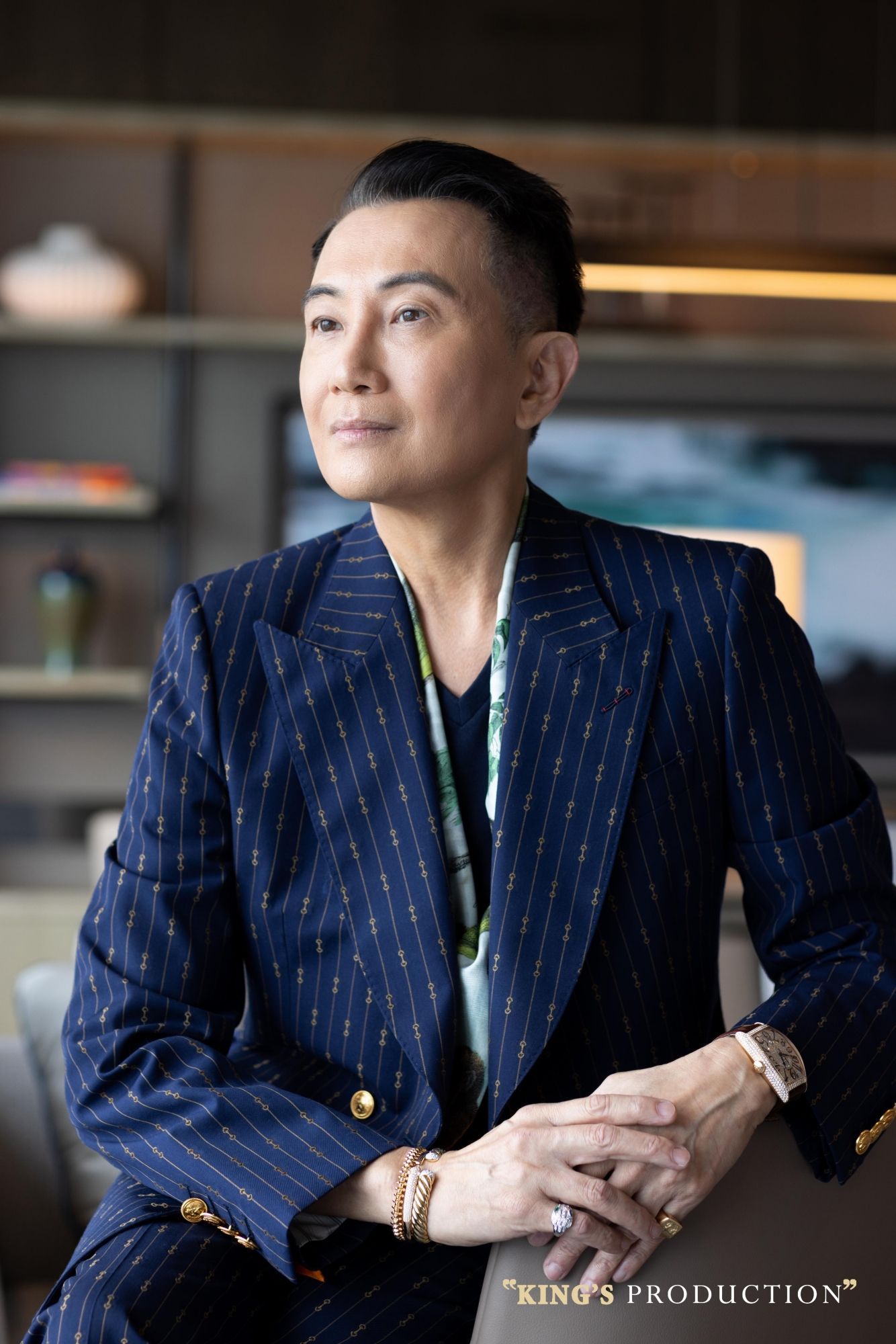 身穿細條紋西裝的男子，斜倚身子，目光移開。室內，背景淡雅，手臂放在椅子上。