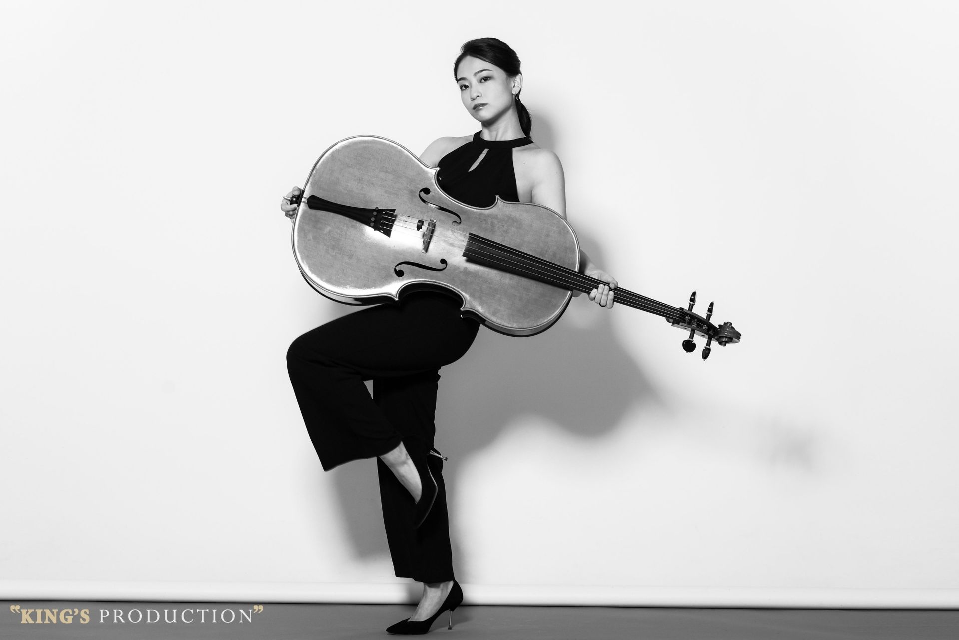 身穿黑色服裝的女子手持一把大型空心大提琴，在白色背景上單腳站立。