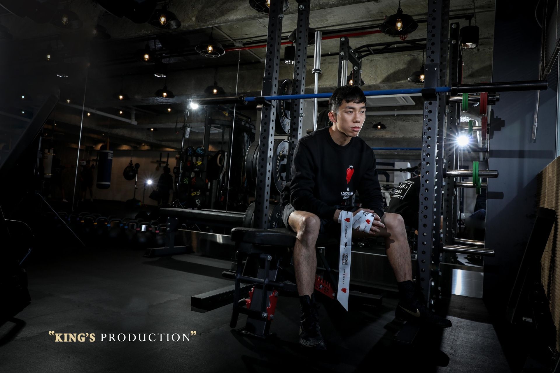 一名男子手持劍，坐在健身房。他身著黑衣，低頭看著前方，手上纏著繃帶。背景昏暗。