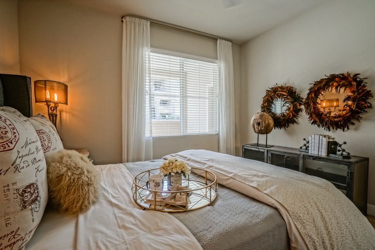 Cozy apartment bedroom with a neatly made bed, window with blinds, sheer white curtains, and decorative mirrors.