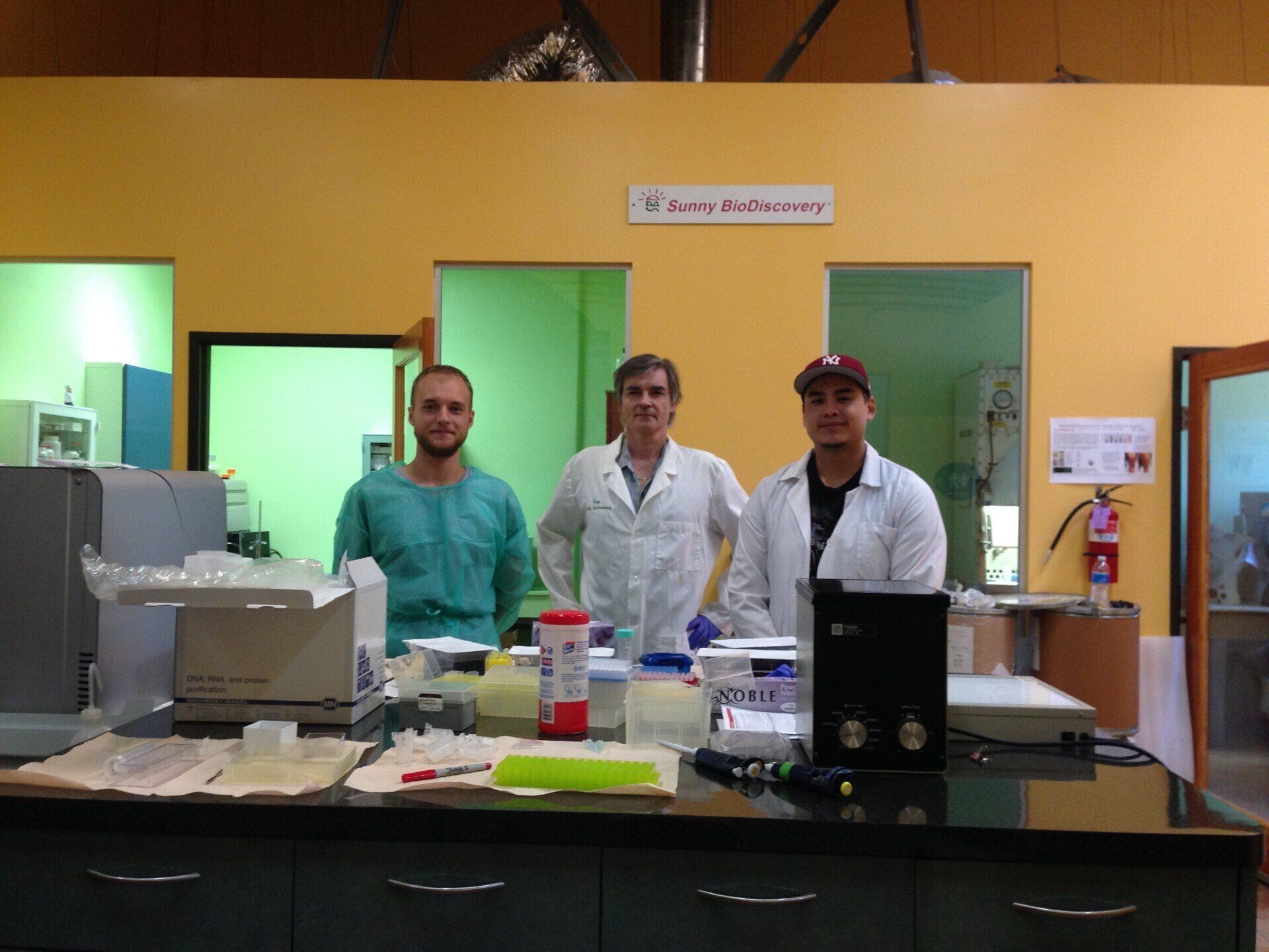 Three men in lab coats standing behind a lab bench. Yellow wall with green doorways.