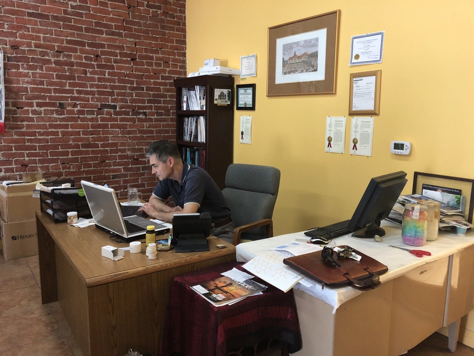 A man works at a desk in an office, surrounded by computers, paperwork, and brick and yellow walls.