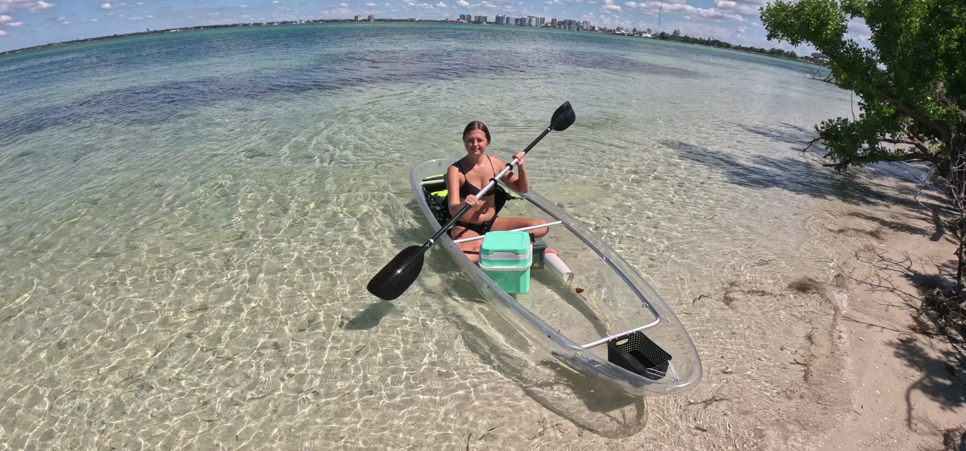 Sarasota skyline, Watersports now kayaking to shore in a clear kayak floating on top of clear water with white sand.