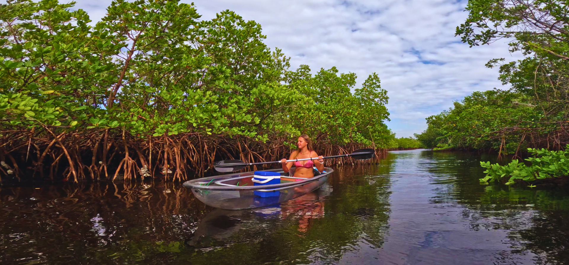 woman kayaking through a mangrove canal full of vibrant green trees with brown roots. The kayak is clear, and has a blue cooler sitting in the bow of the boat.