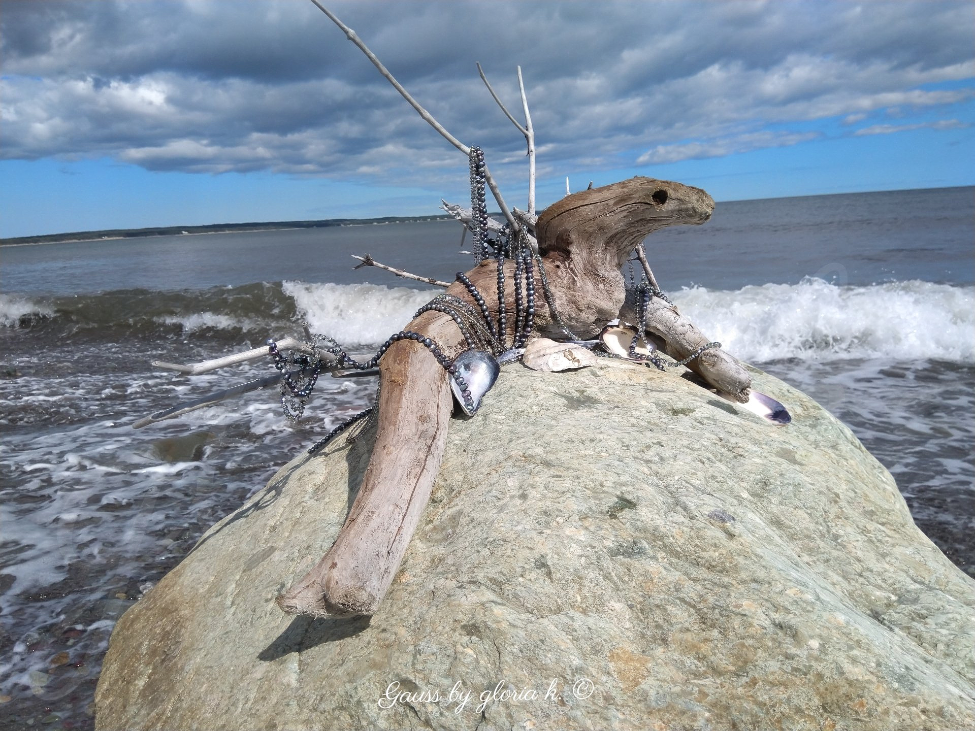 Driftwood sea creature draped with peacock pearls and crystals on sea boulder.