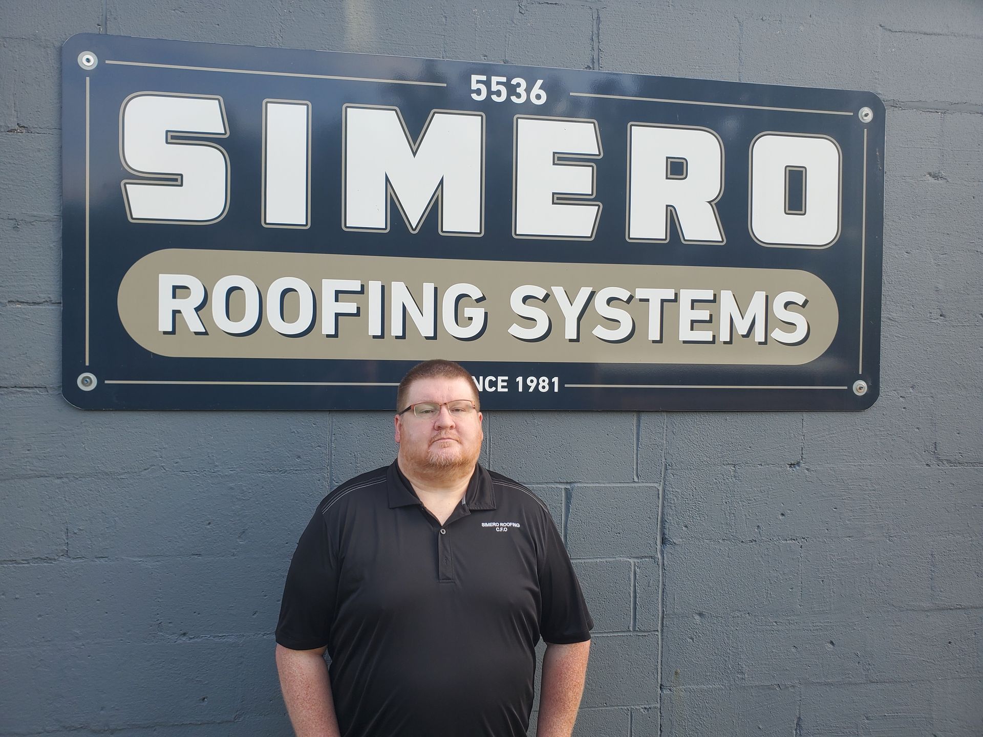 Man standing in front of Simero Roofing Systems sign. The sign is dark blue and gold, brick wall background.
