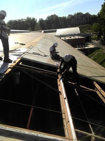 Construction workers on a roof, removing old materials. They wear hard hats and safety harnesses in the sunlight.