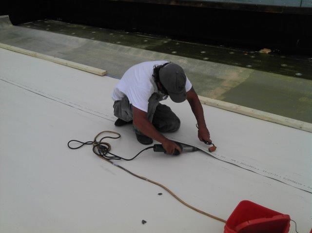 Person kneeling on a white surface, using a power tool, likely repairing a roof.