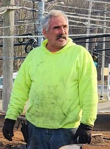 Man in a neon green hoodie, black gloves, and jeans, looking to the side outdoors, possibly working.