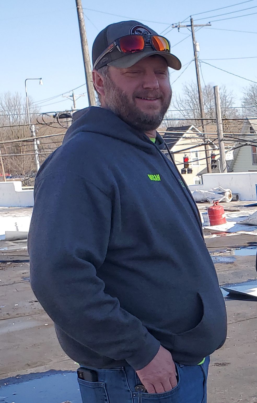Man in cap and hoodie smiles outside, arms relaxed, near a construction site.