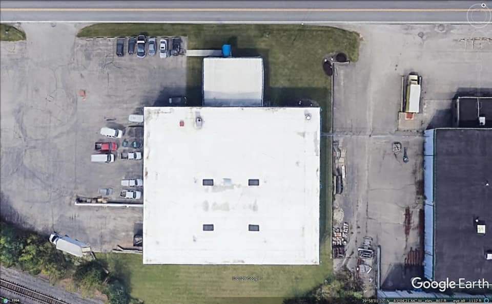Overhead view of a white-roofed industrial building, surrounded by parking, roadways, and other structures.