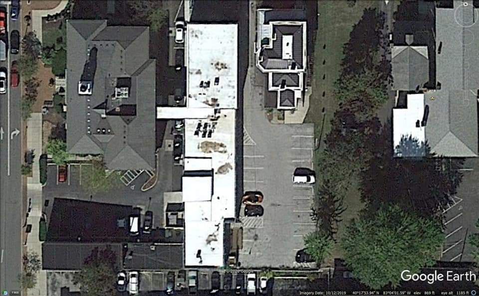 Aerial view of several buildings, a parking lot, and trees. White and gray roofs dominate.