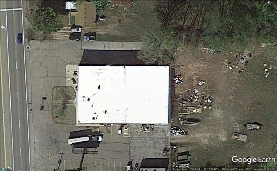 Overhead view of a white-roofed building and a parking area, surrounded by vegetation and a street.