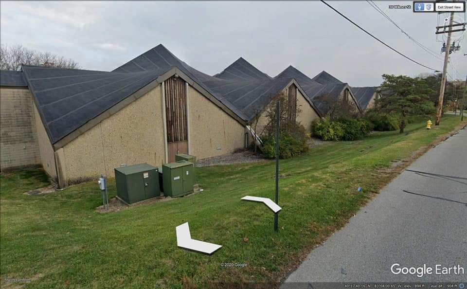 Tan building with angular, black roofs; green grass and a street in front.