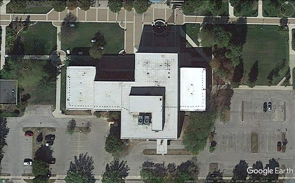 Aerial view of a building with a flat roof, surrounded by trees, parking lots, and walkways.