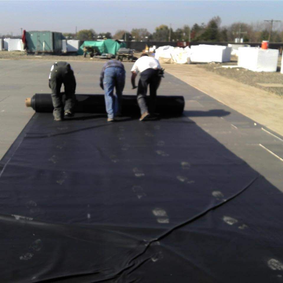 Three workers unrolling a large black tarp on an outdoor surface.