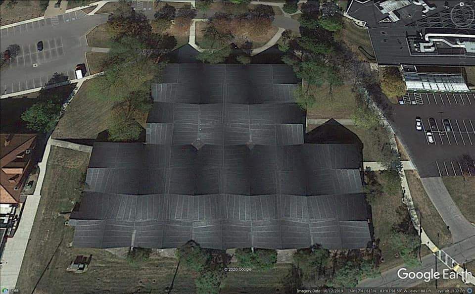 Aerial view of a large, dark-roofed building with a unique tiered structure, surrounded by trees and parking lots.