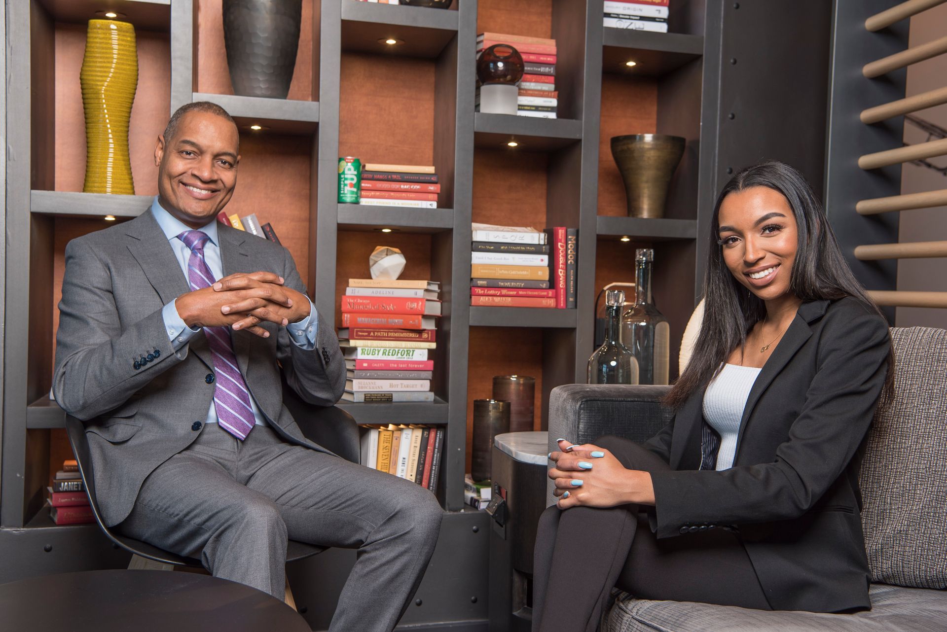 A man and a woman are sitting on a couch in front of a bookshelf.