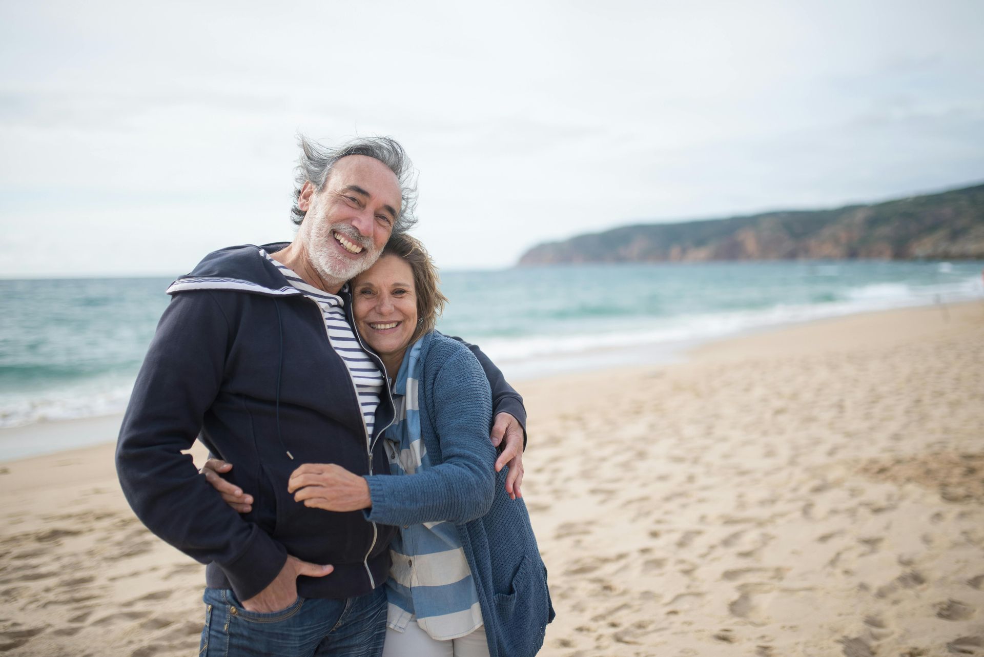 Couple embracing on a sandy beach, smiling. Ocean and cliffs in background.