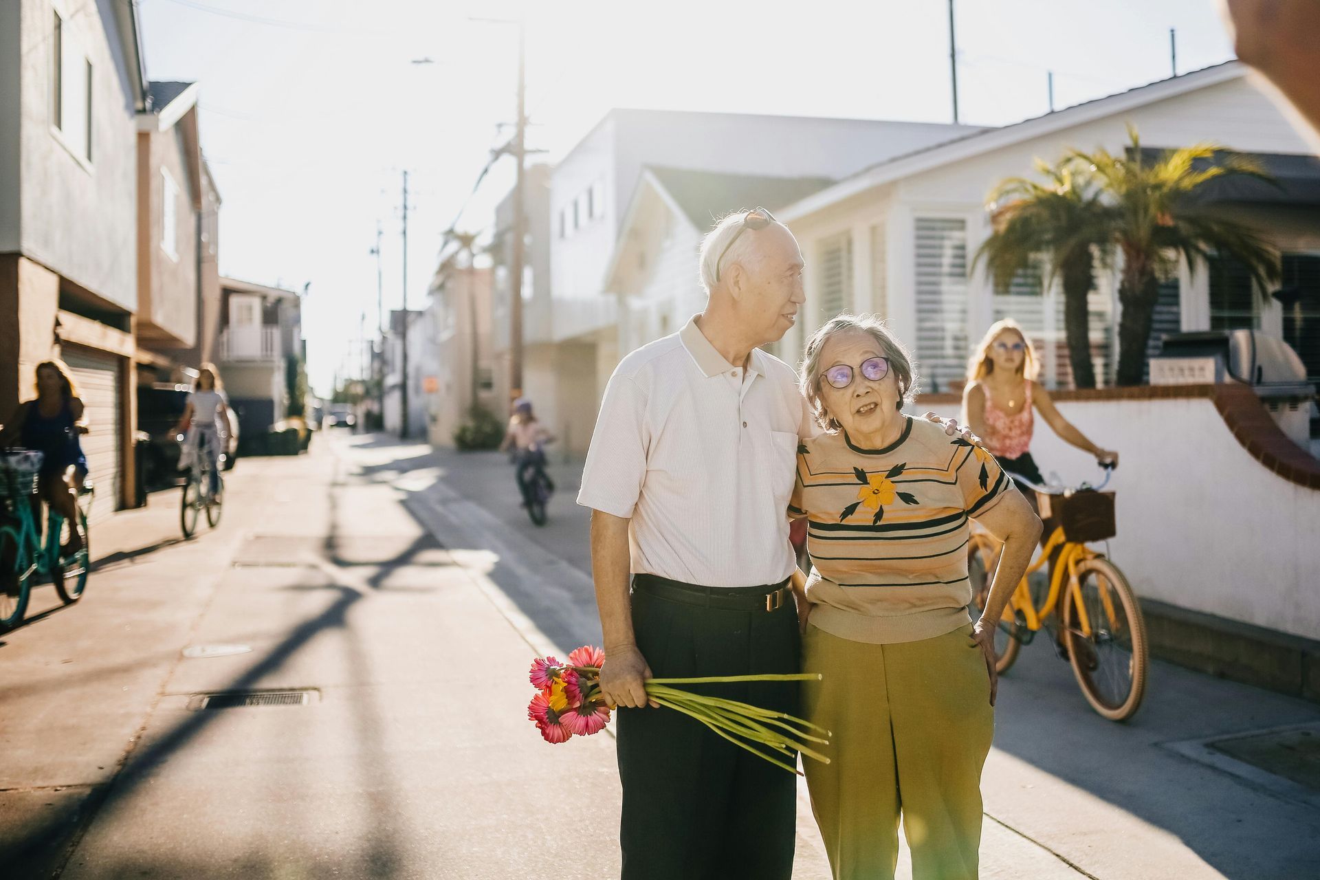 Elderly couple walking, embracing, holding flowers on a sunny street; cyclists nearby.