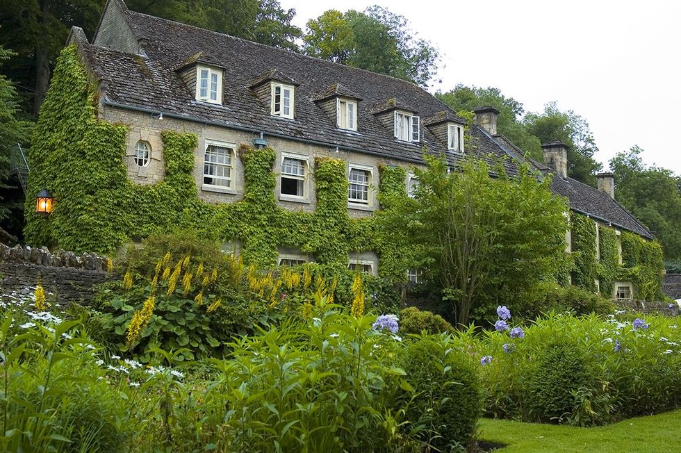 Stone building covered in ivy with a lush garden, set in a natural, green landscape.